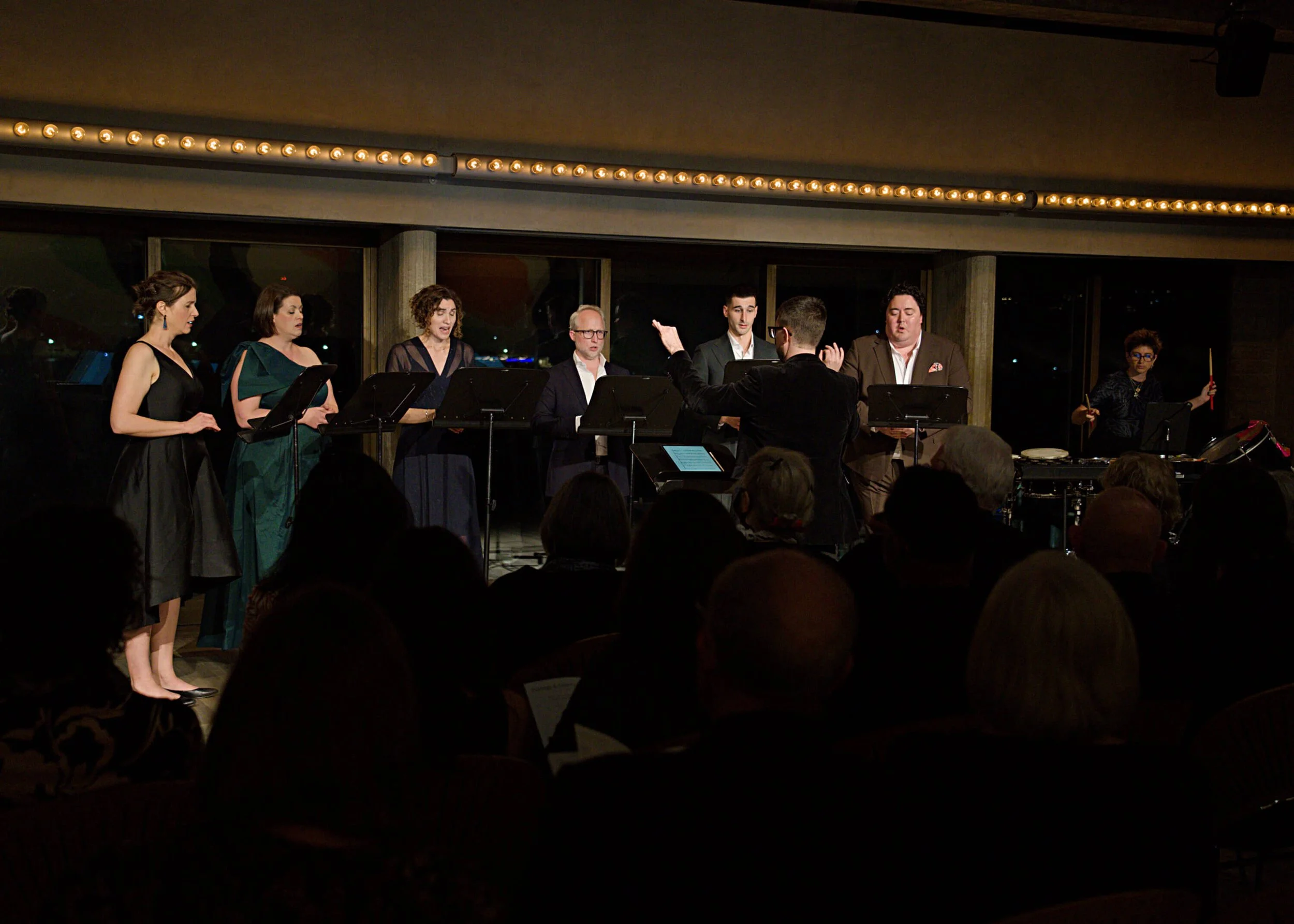 A choir performing on stage with a conductor guiding them. Women are singing and dressed in formal attire, with a musician playing drums on the right. Audience members are visible in the foreground.