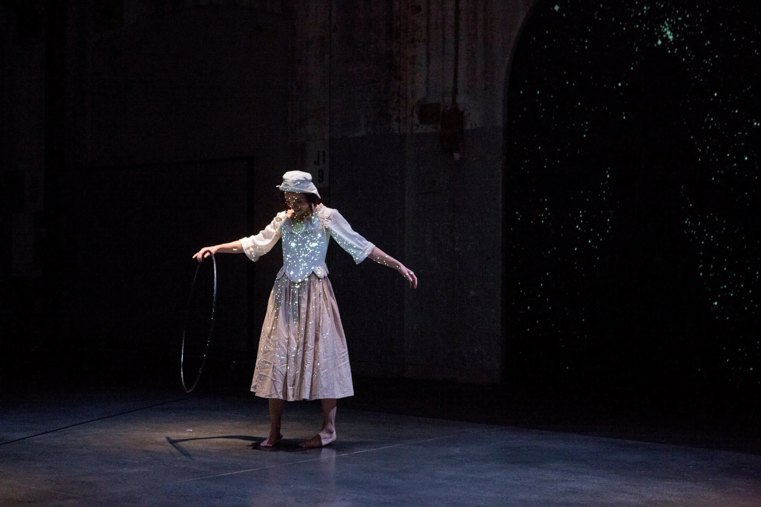 A performer wearing a vintage-style ensemble, including a white blouse, long skirt, and hat, on stage with a dark background and specks of light projected onto her and the backdrop.