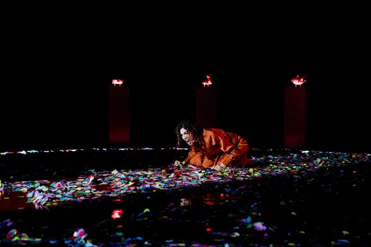 Jessica O'Donoghue in an red dress crouches on a dark stage, surrounded by colorful confetti, with three small red-lit sculptures of animals on red pedestals in the background.