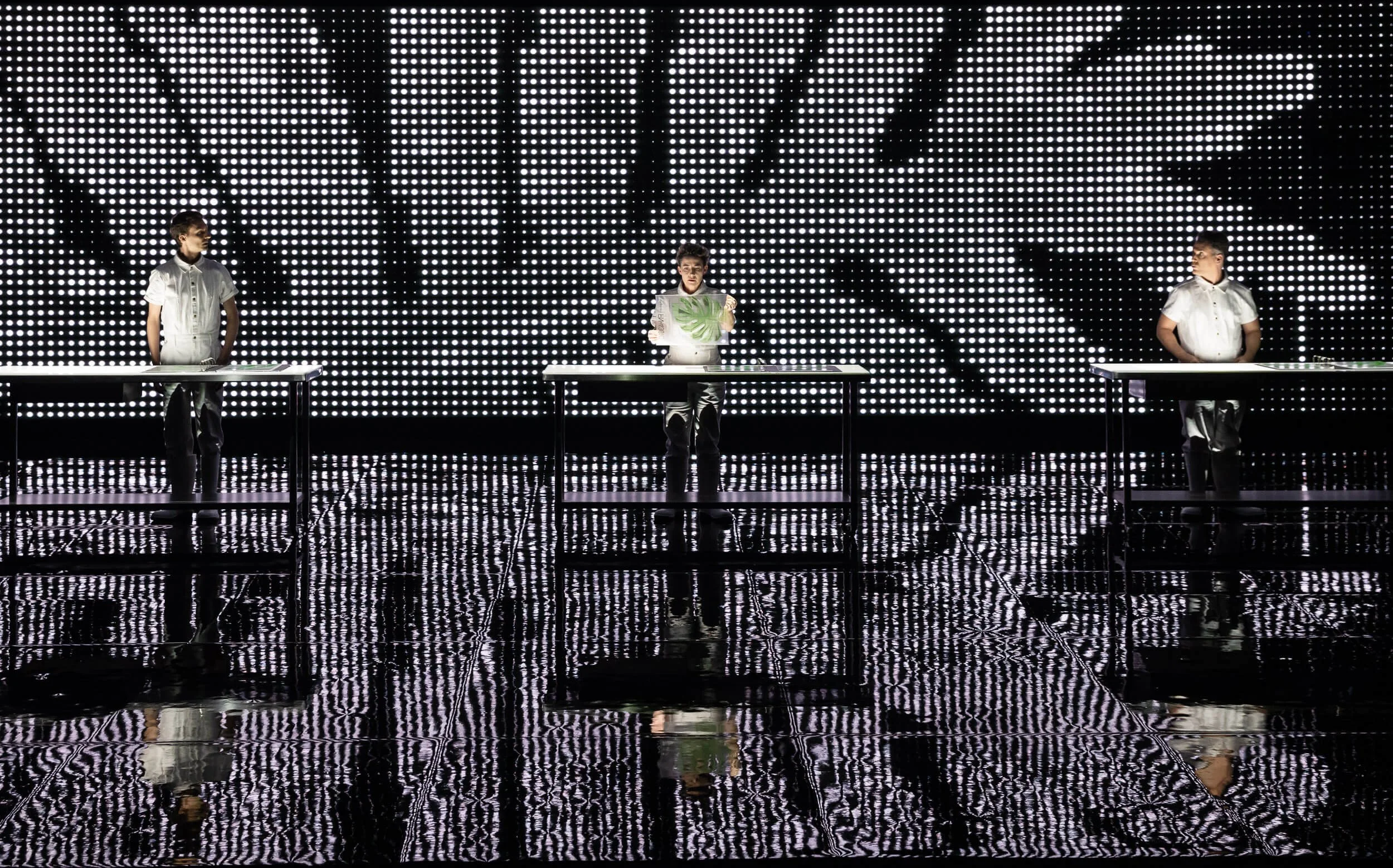 Three people standing at separate tables in front of a large LED wall displaying the words "RAVE LIKE".