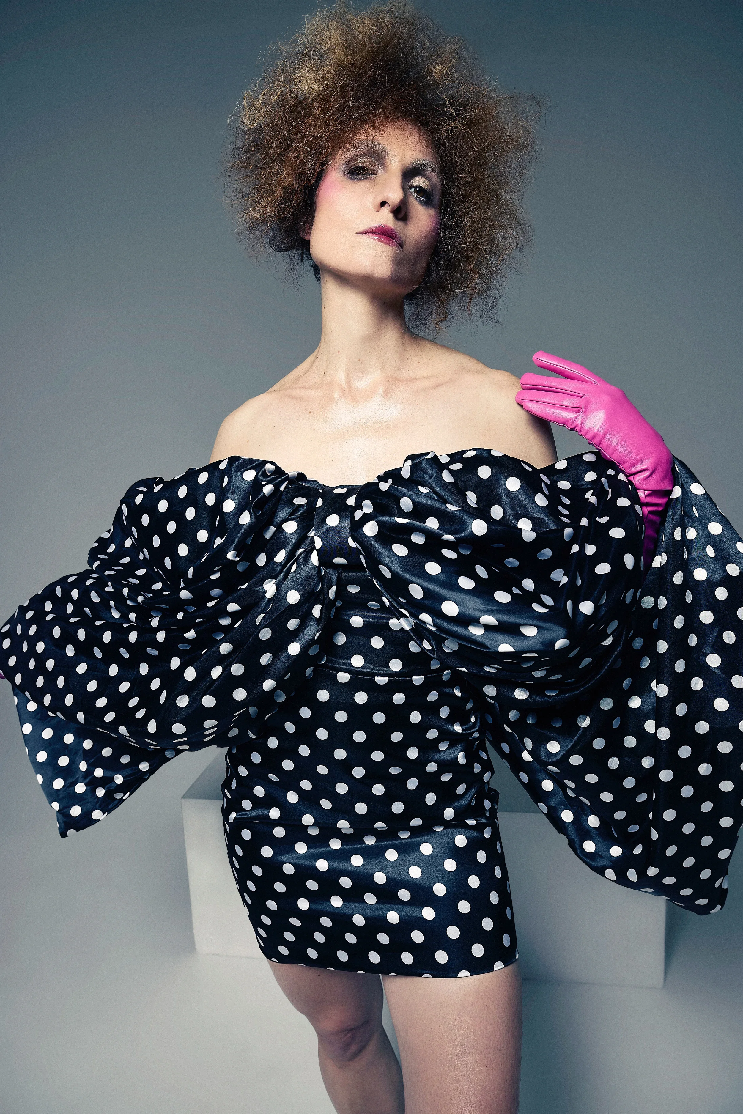 Jessica O'Donoghue with curly hair wearing a black dress with white polka dots, pink gloves, and bold makeup.