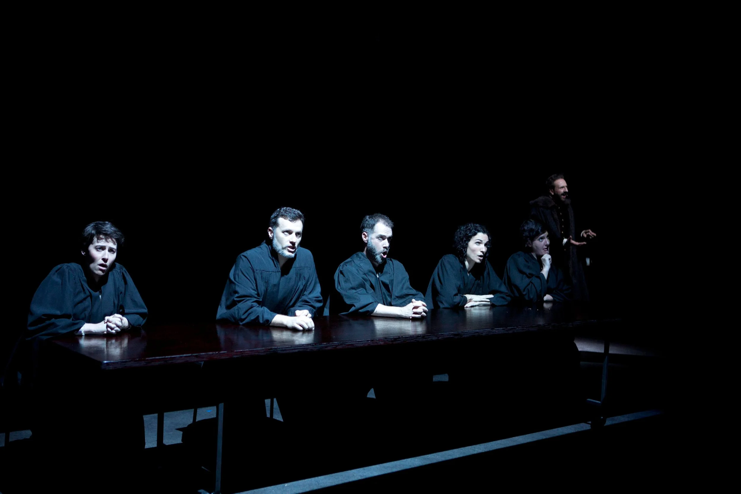 Six people dressed in black robes sitting at a dark table with a man standing at the far right, all appearing to be in a theatrical or performance setting.