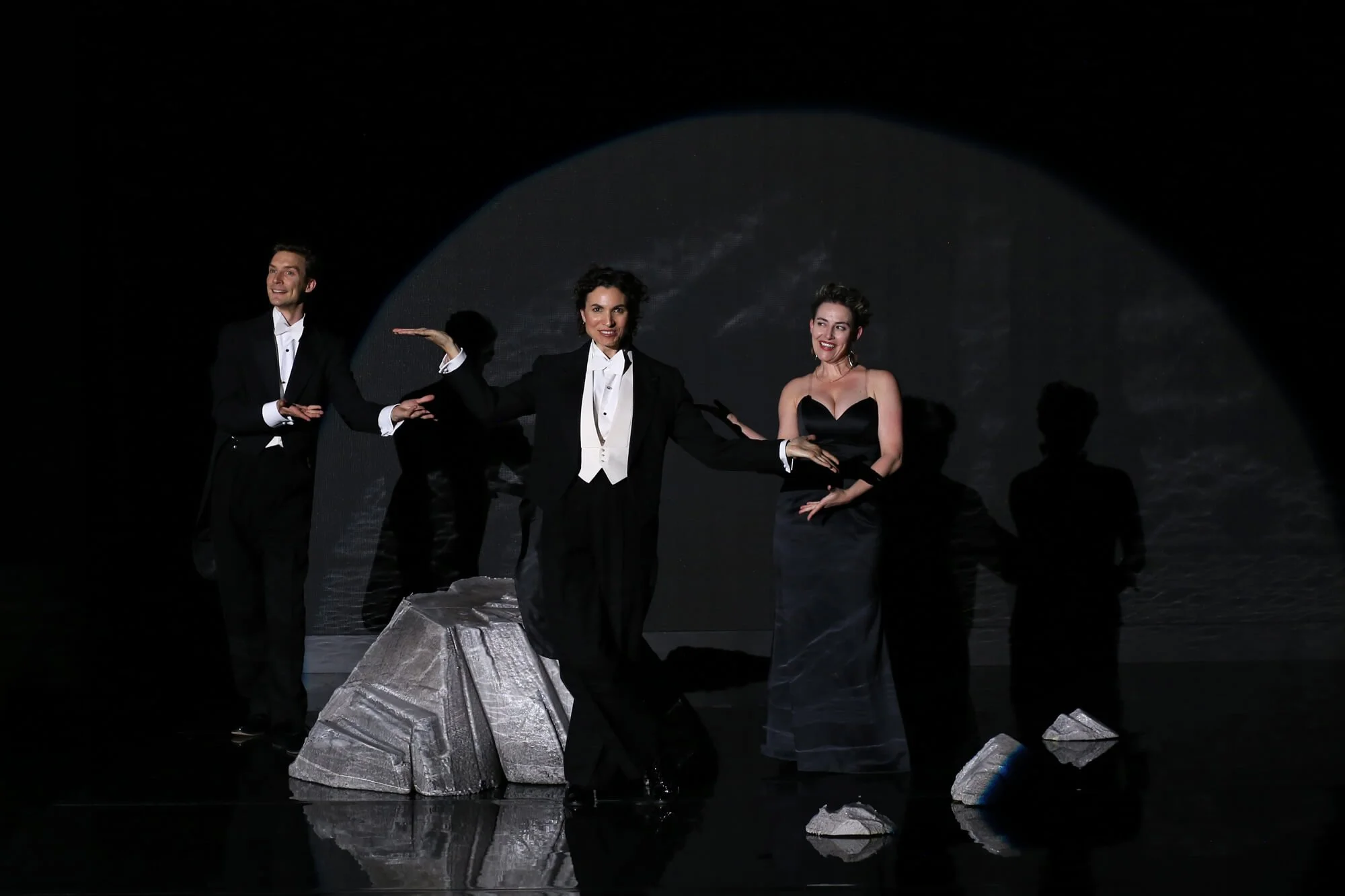 Group of performers dressed in formal attire on a stage with rocks and a celestial background.