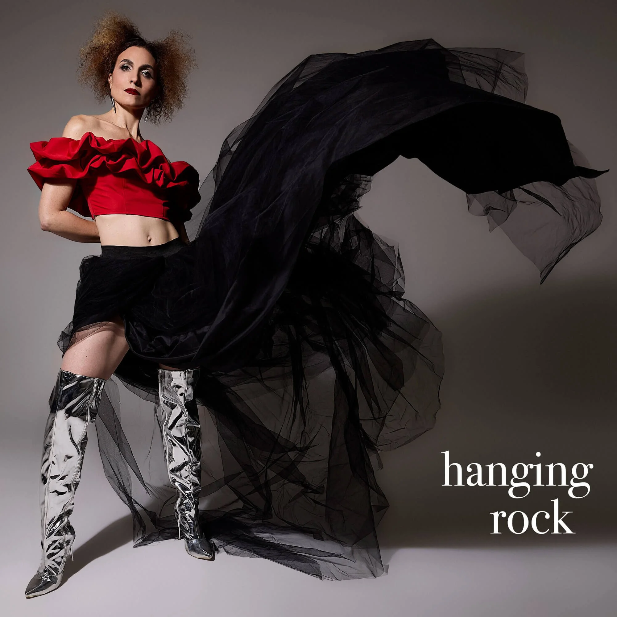 Fashionably dressed Jessica O'Donoghue with curly hair, wearing a red ruffled crop top, shiny silver pants, and metallic boots, standing next to black tulle fabric.