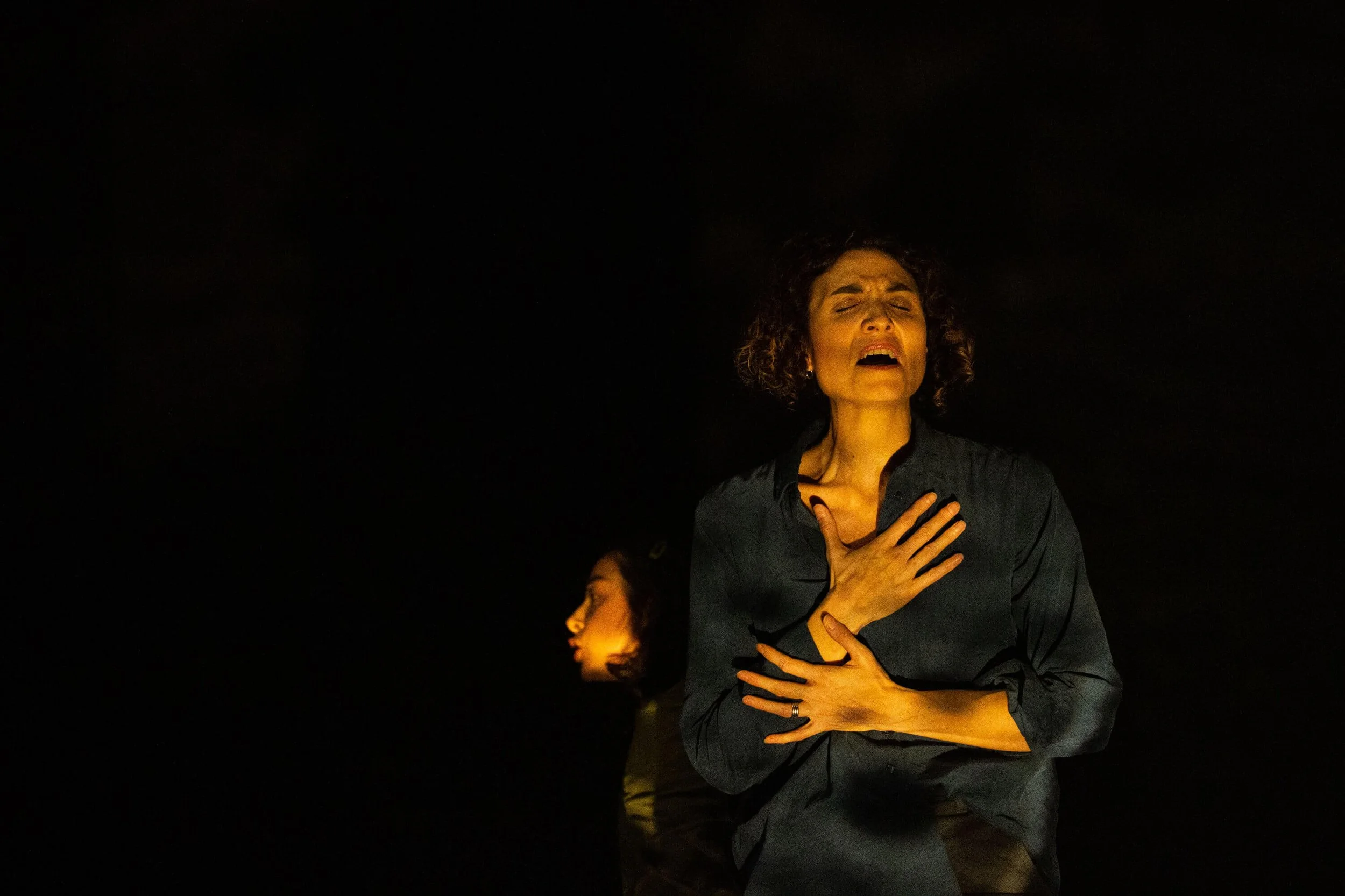 Jessica O'Donoghue with curly hair, wearing a dark shirt, with her eyes closed and mouth open, appears to be emotional or singing, with one hand on her chest and the other hand on her stomach, in a dark setting with faint light.
