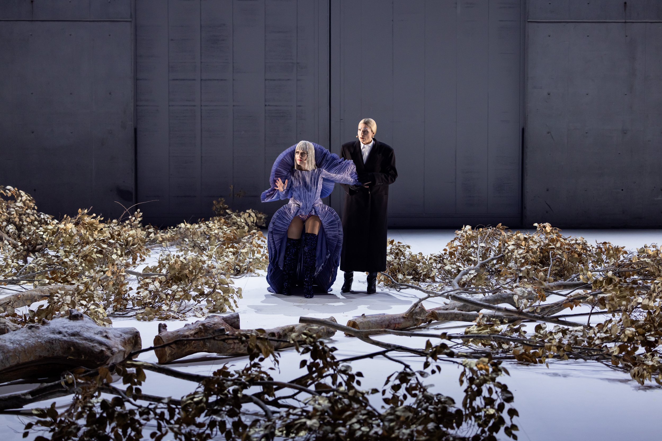 Two performers on stage surrounded by dried leaves and branches, minimalist gray wall background, one standing and one seated in elaborate costumes.