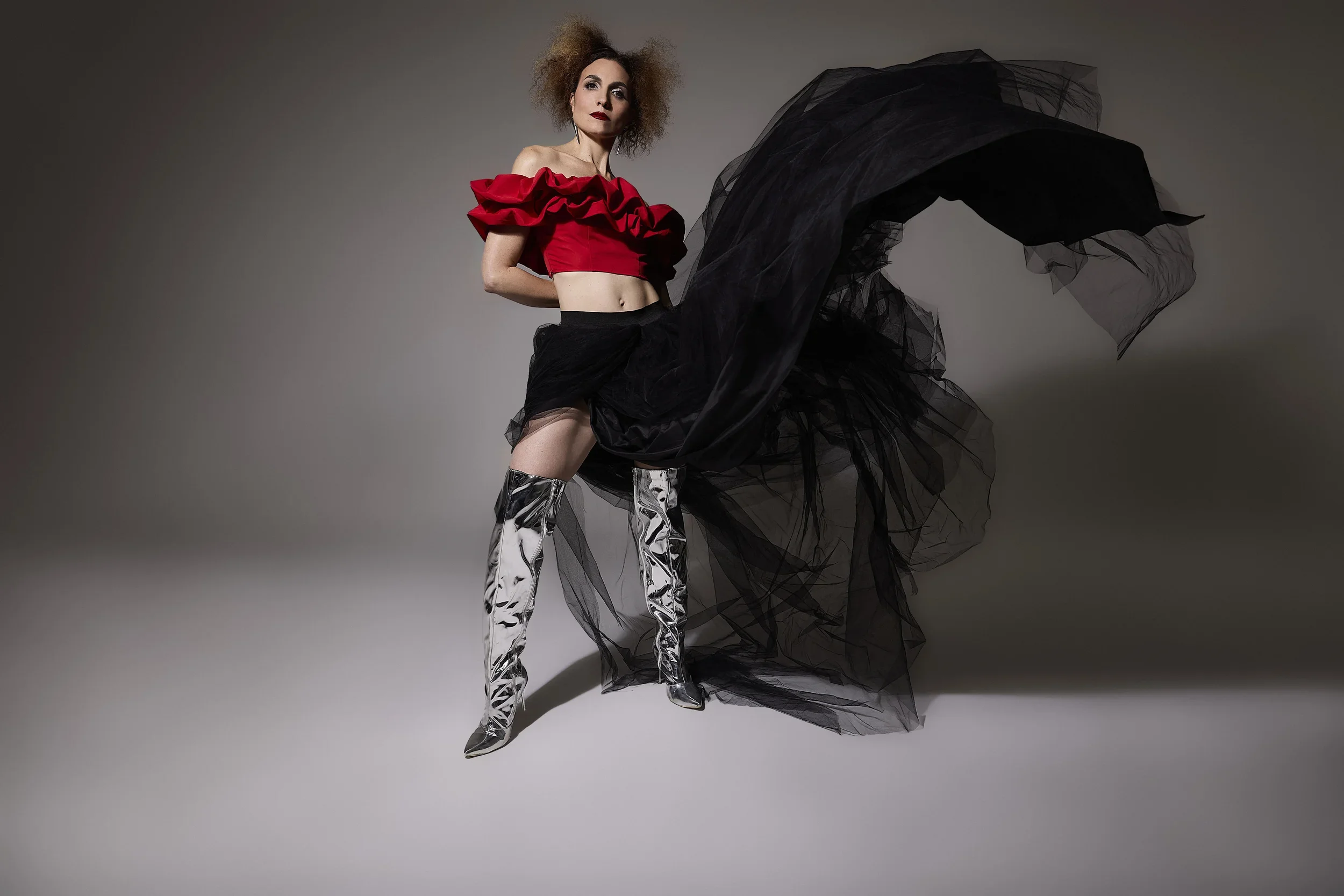 Jessica O'Donoghue with curly hair in a red off-shoulder top with ruffles, black tulle skirt, and shiny silver thigh-high boots standing against a plain background.