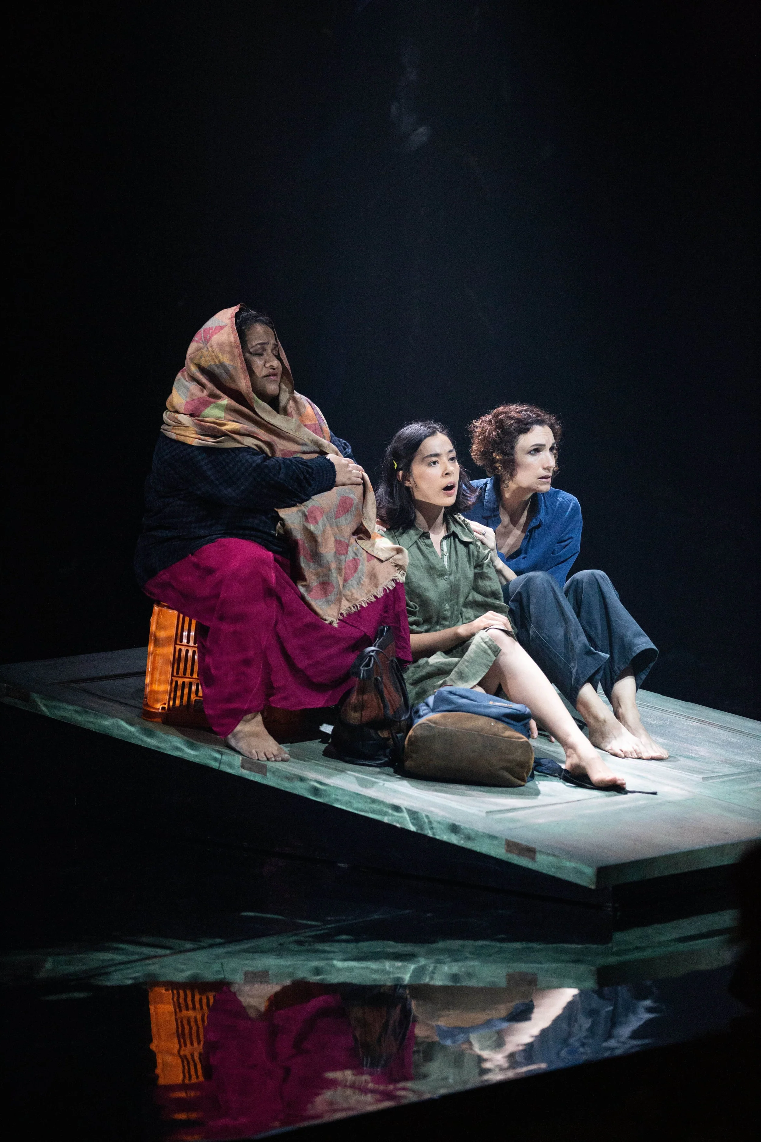 Three women sitting on a floating platform, characters from a theatrical performance, with various bags and personal items around them, set against a black background.