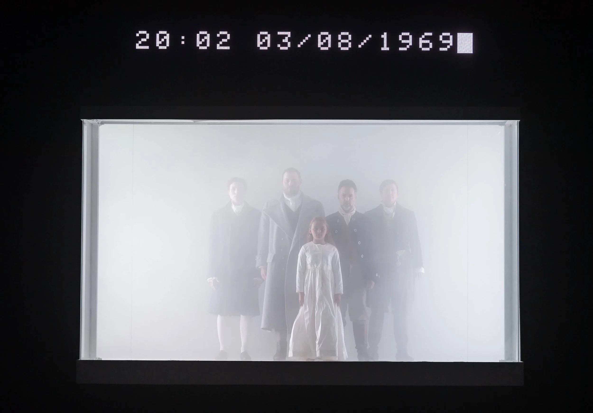A family of six people, including children, standing in an elevator with a bright, white interior. The elevator has a digital display above it showing the date 20/02/03 and the year 1969.