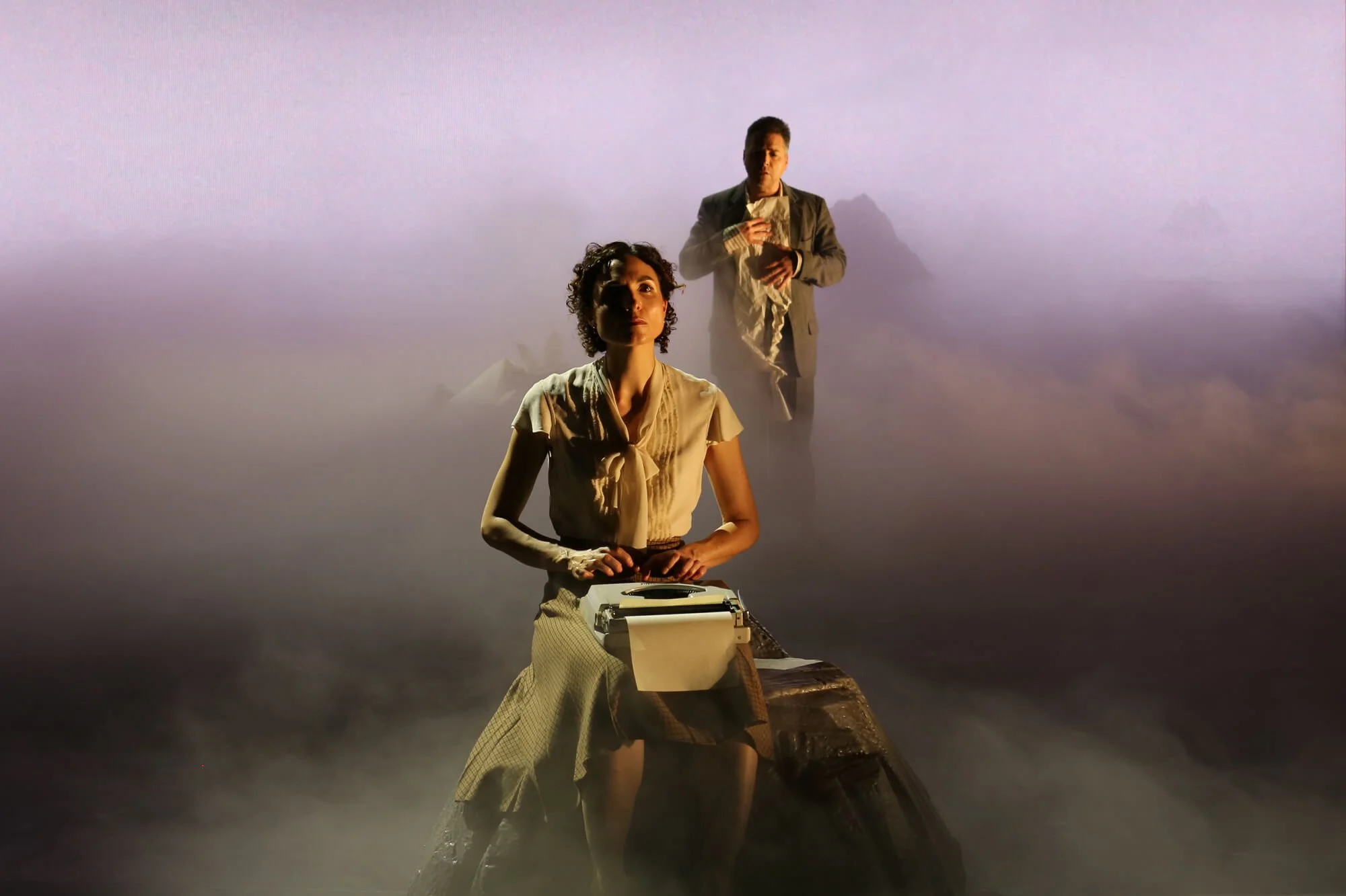 Jessica O'Donoghue with short curly hair wearing a beige blouse and beige skirt standing on a platform with a typewriter in front of her, and a man in a suit standing behind her holding a paper, in a foggy, surreal setting.
