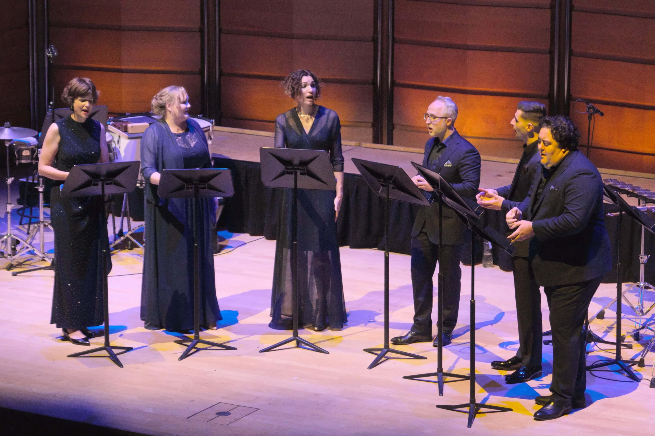 Group of six musicians singing together on stage, with music stands and instruments around them.