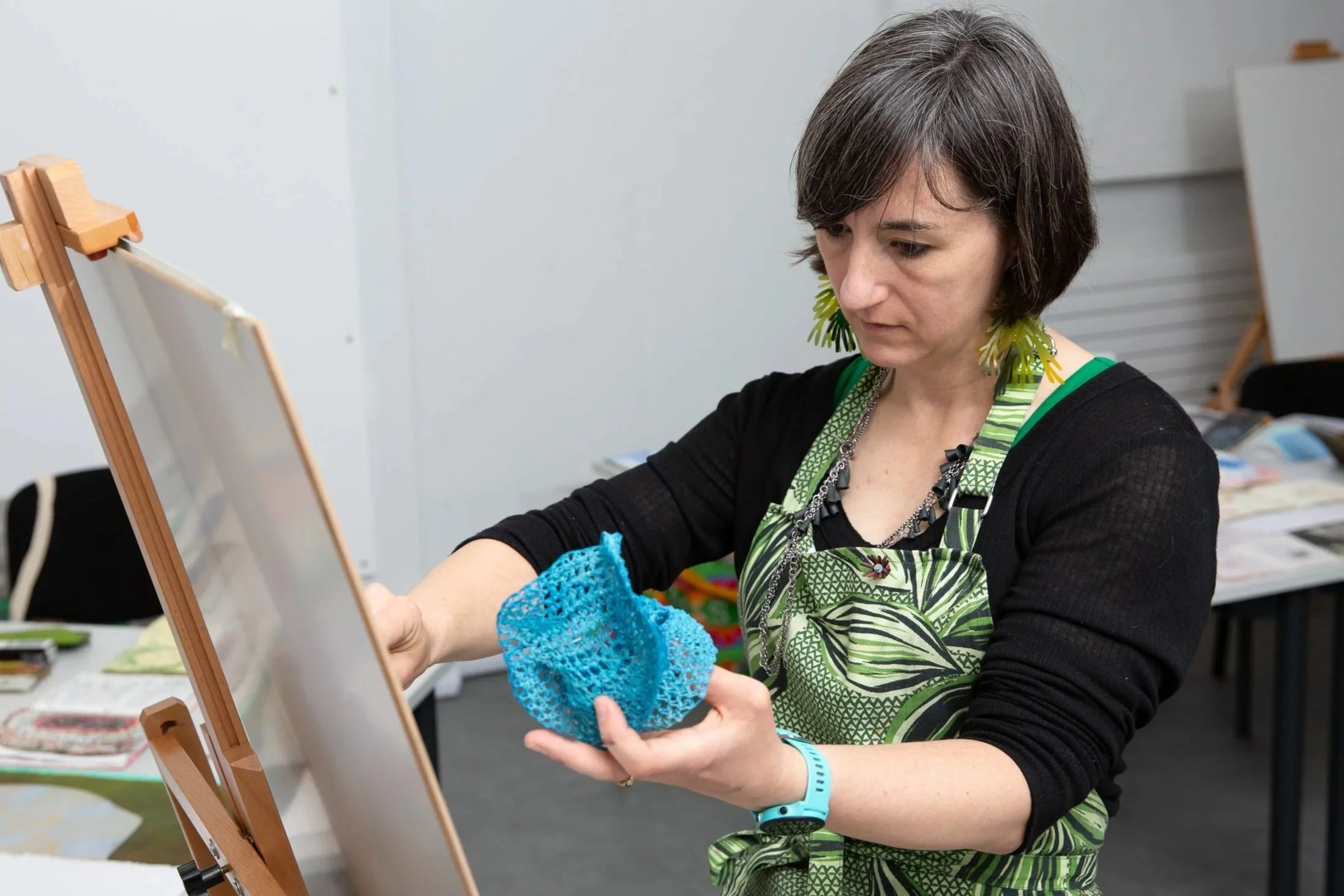 Woman in a green and black apron using a blue knitting loom to create fabric, standing in front of a white wall with a wooden easel in the background.