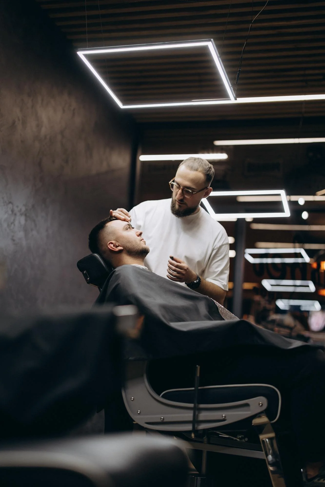 A man receiving a haircut from a barber in a modern barber shop, with neon lighting fixtures on the ceiling.