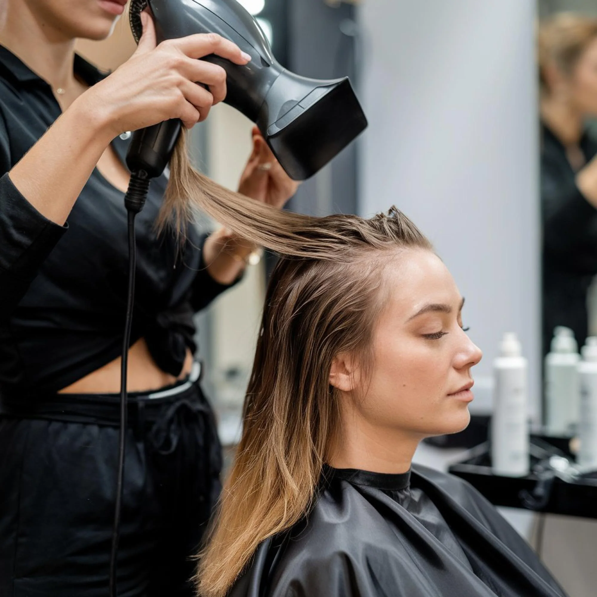 A woman getting her hair blow-dried at a salon. She is sitting with her eyes closed, and a stylist is holding a hairdryer directed at her hair.