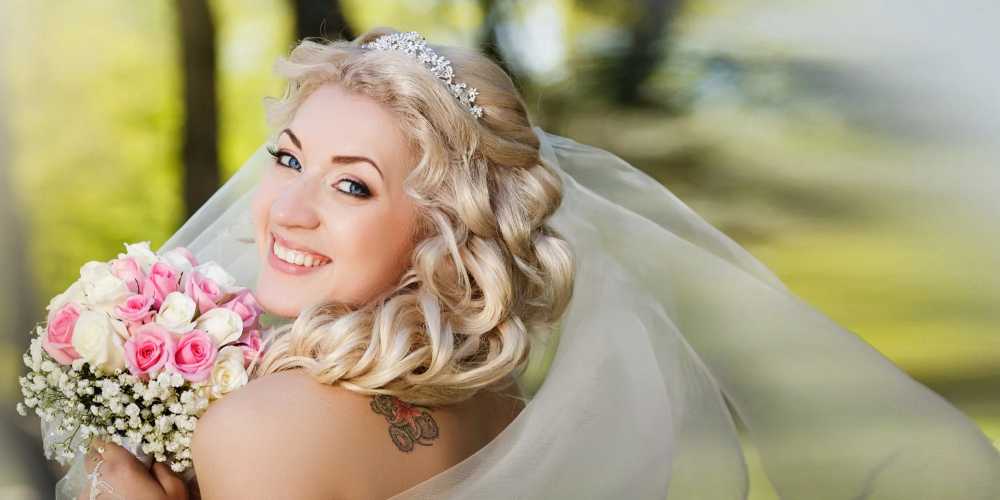 A smiling bride with blonde curly hair and blue eyes, wearing a tiara and veil, holds a bouquet of pink and white roses with baby's breath, outdoors with blurred green trees in the background.