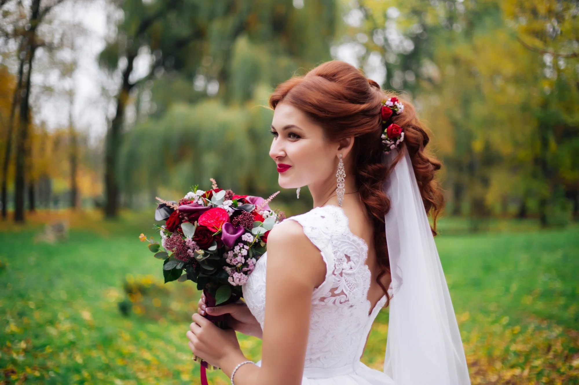 A bride with red hair, wearing a white lace wedding dress and large earrings, is holding a bouquet of red, pink, and purple flowers with greenery, in a park with green trees and yellow leaves, during daytime.
