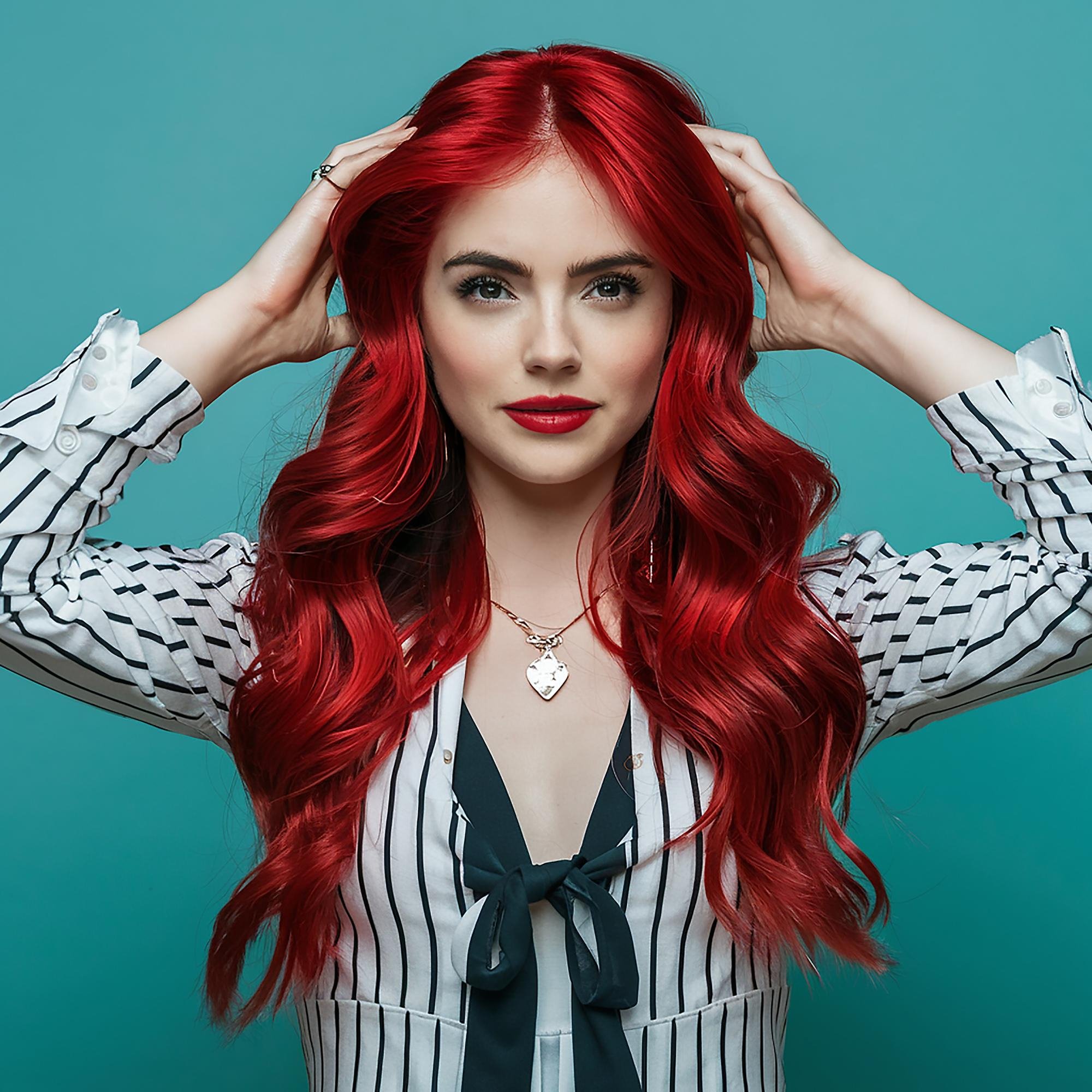 A woman with long, vibrant red hair styling her hair with both hands, wearing a black and white striped blouse, a black bow tie, and a silver necklace with a heart pendant. She stands against a teal background.