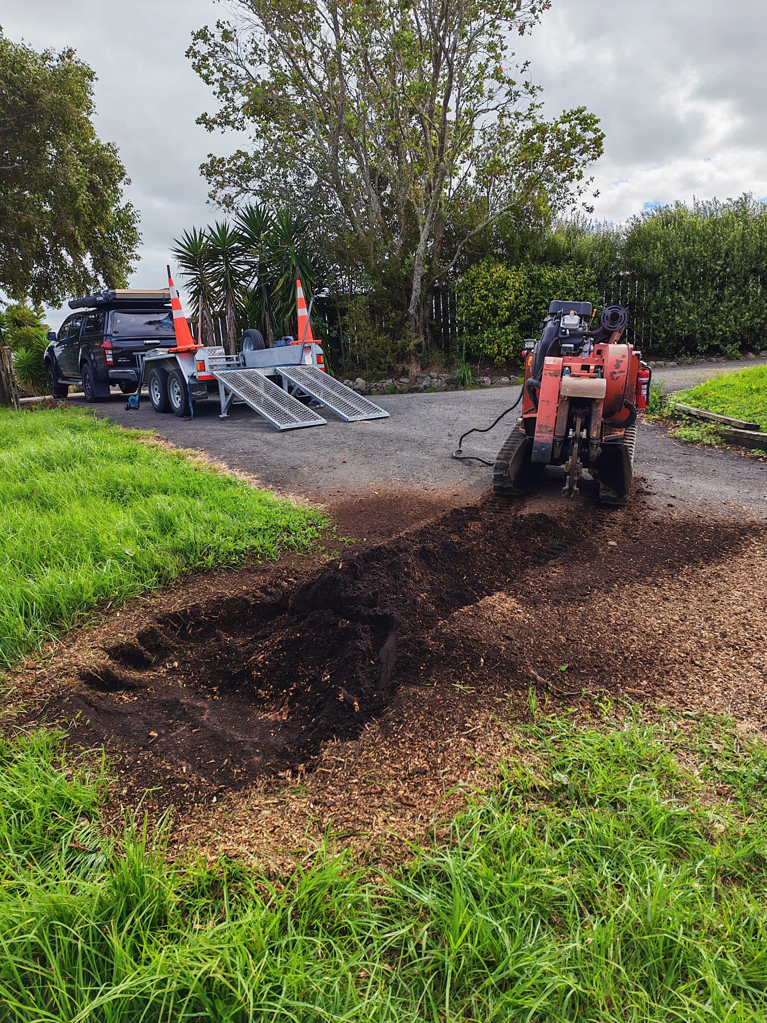 A small excavator digging a hole in a grassy area with a black pickup truck in the background, nearby trees and bushes, cloudy sky overhead.