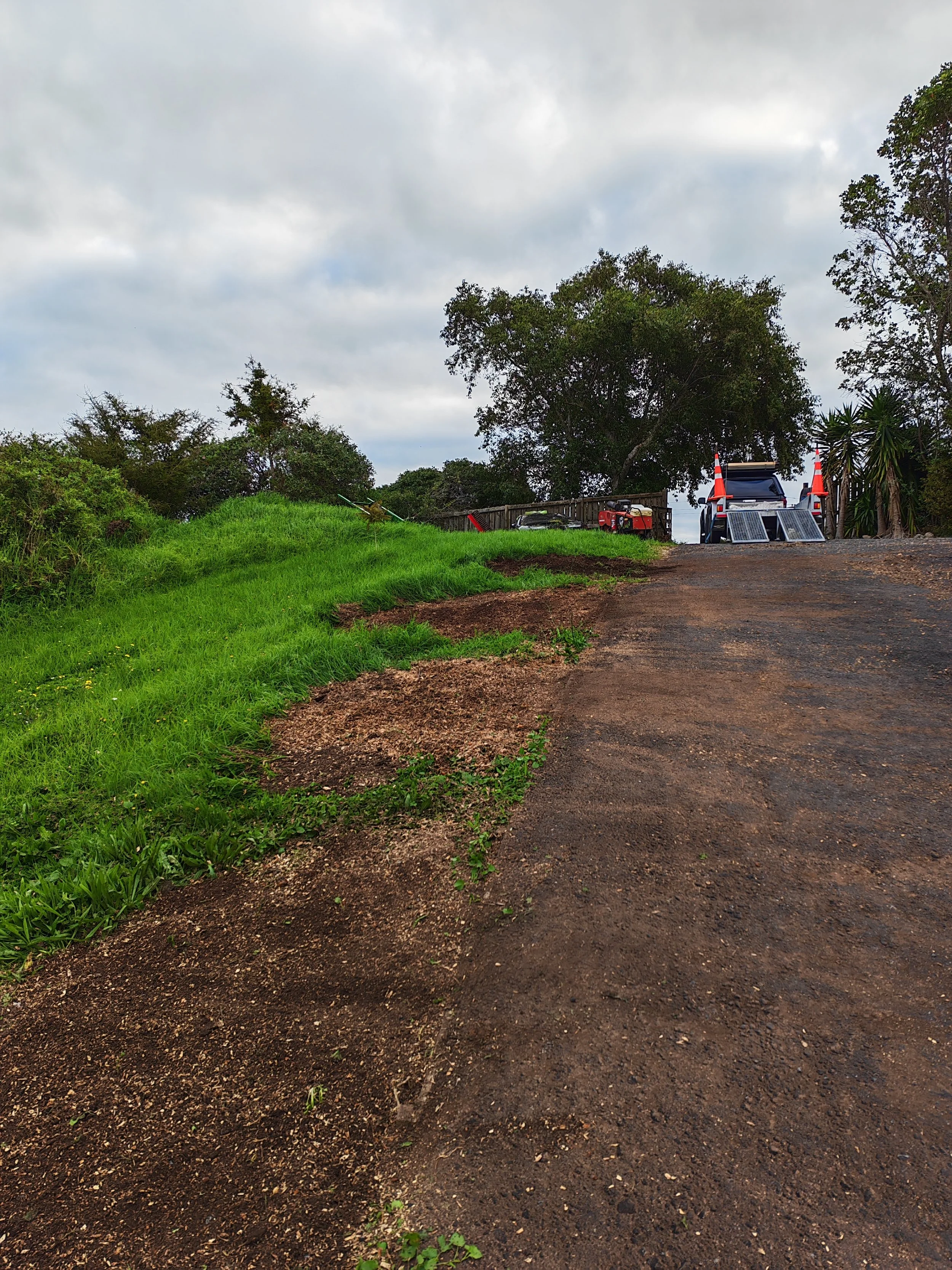 A dirt driveway or path curves to the right, with green grass and plants on the left side. There are trees and a wooden fence at the end of the driveway. Two orange traffic cones and a barricade block the road, and a vehicle is parked behind them, pa