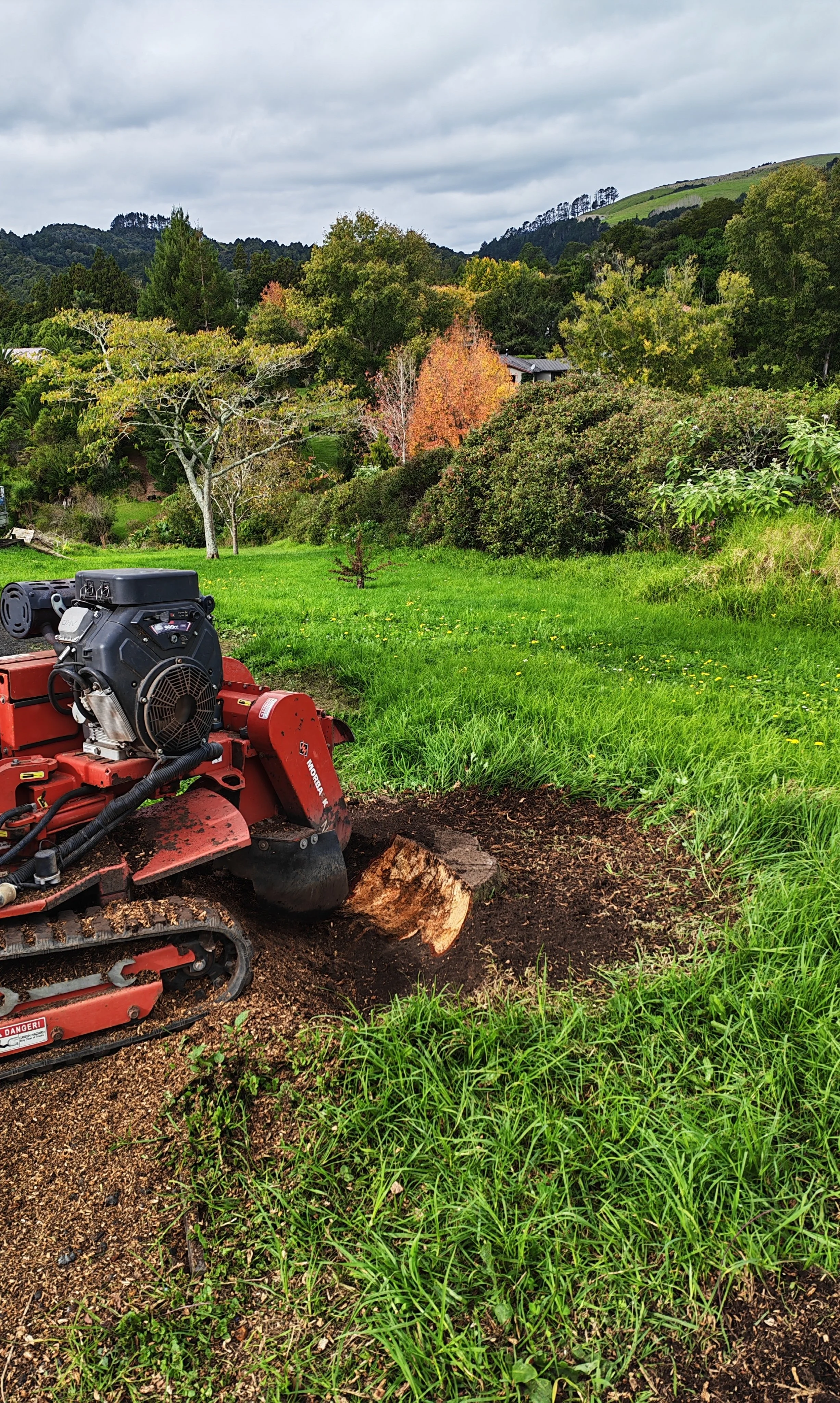Lawn mower working on a patch of dirt and grass, with a scenic background of trees and rolling hills under a cloudy sky.