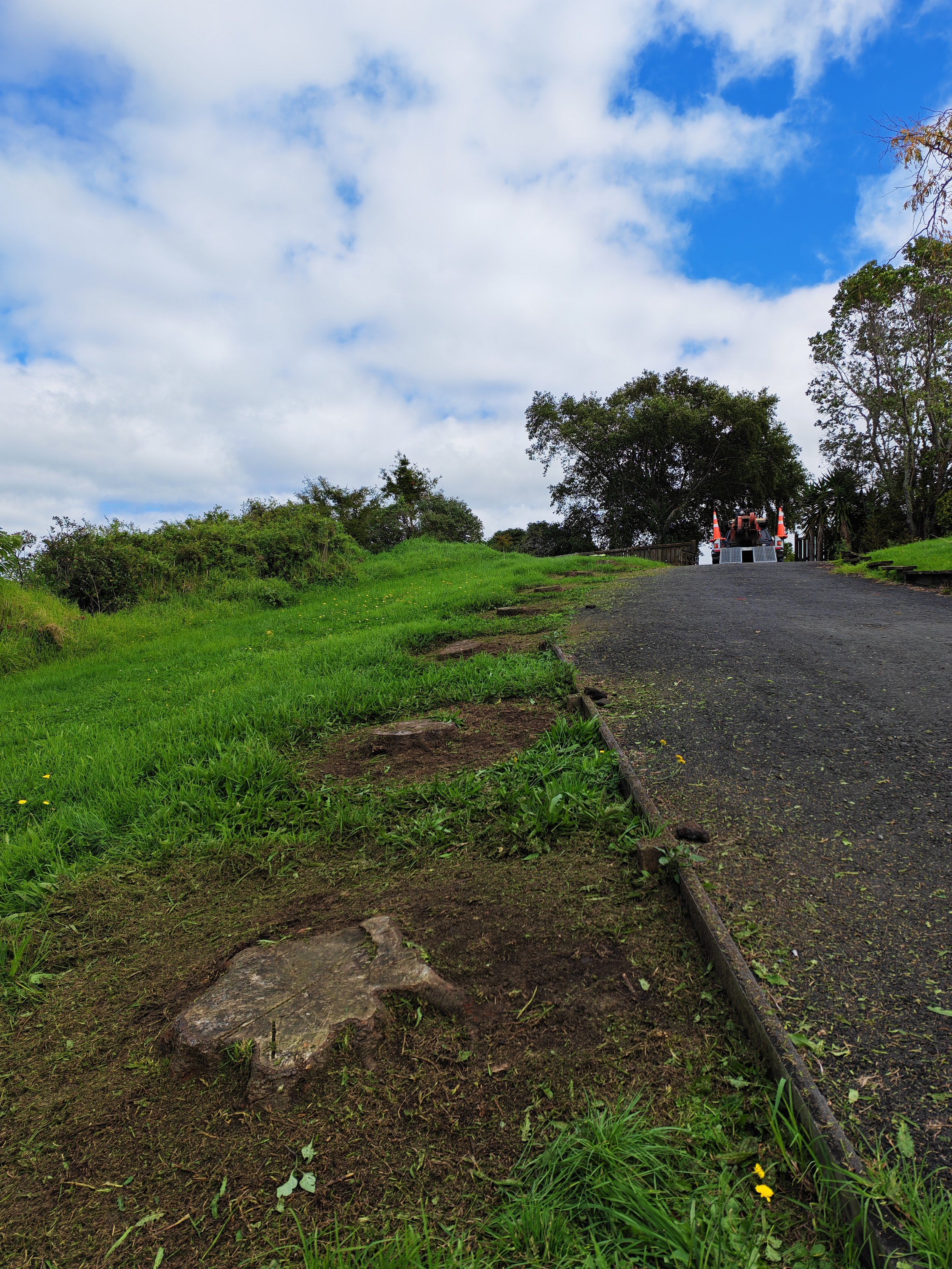 A dirt path with a rusty rail runs through a grassy area with shrubs and trees on either side. In the distance, a red tractor with orange safety cones is parked on the path. The sky overhead is partly cloudy with patches of blue.