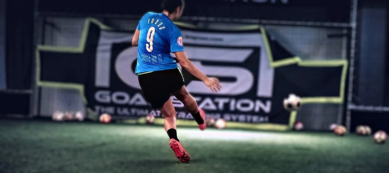 A female soccer player kicking a ball in an indoor training facility with multiple soccer balls and goal nets in the background.