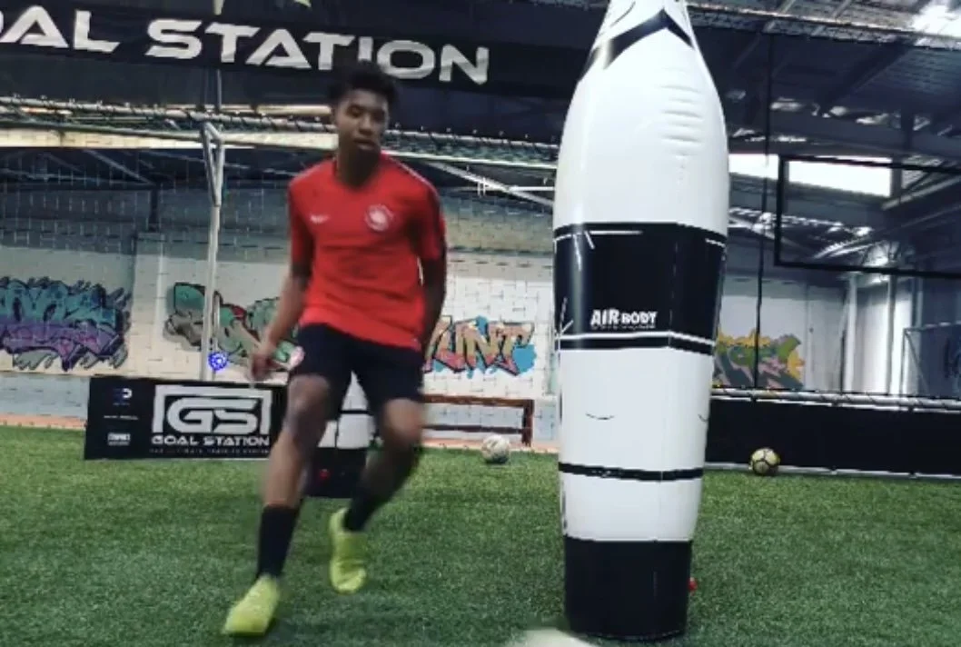 Young boy in red shirt and black shorts training with a punching bag at an indoor sports facility with graffiti on the wall.