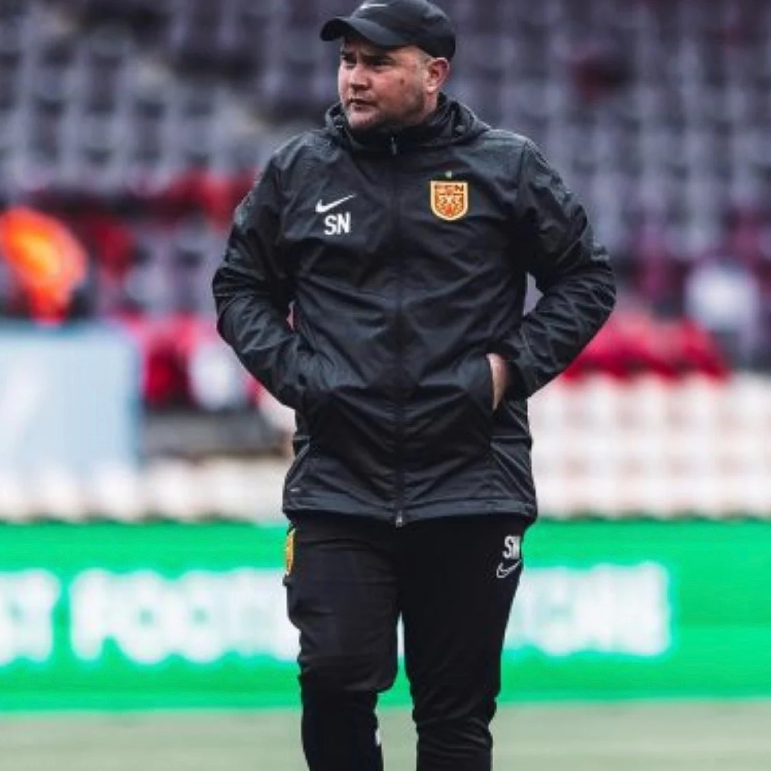 A man dressed in a black Nike sports jacket and pants standing on a sports field with his hands in his pockets, wearing a black cap, with a blurred background of stadium seats.