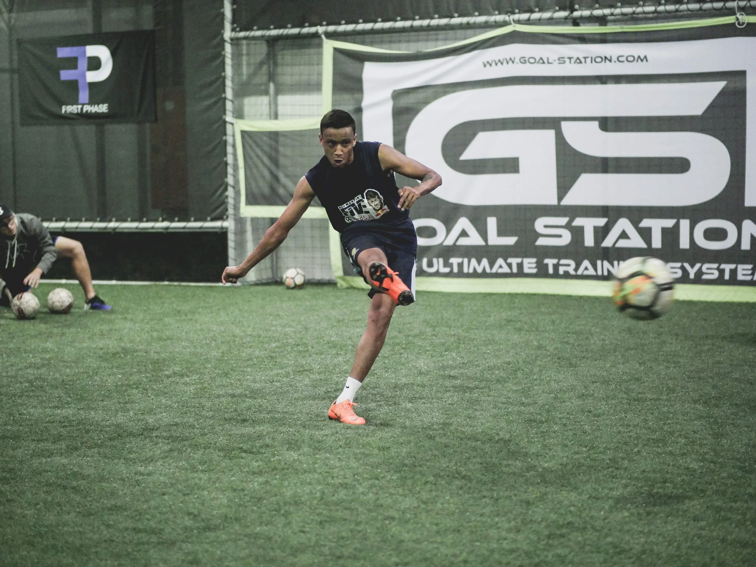 A man in a black sleeveless shirt and shorts playing soccer indoors, kicking a soccer ball with orange cleats on a green turf field amid practice balls and a netted wall with Goal Station banners.