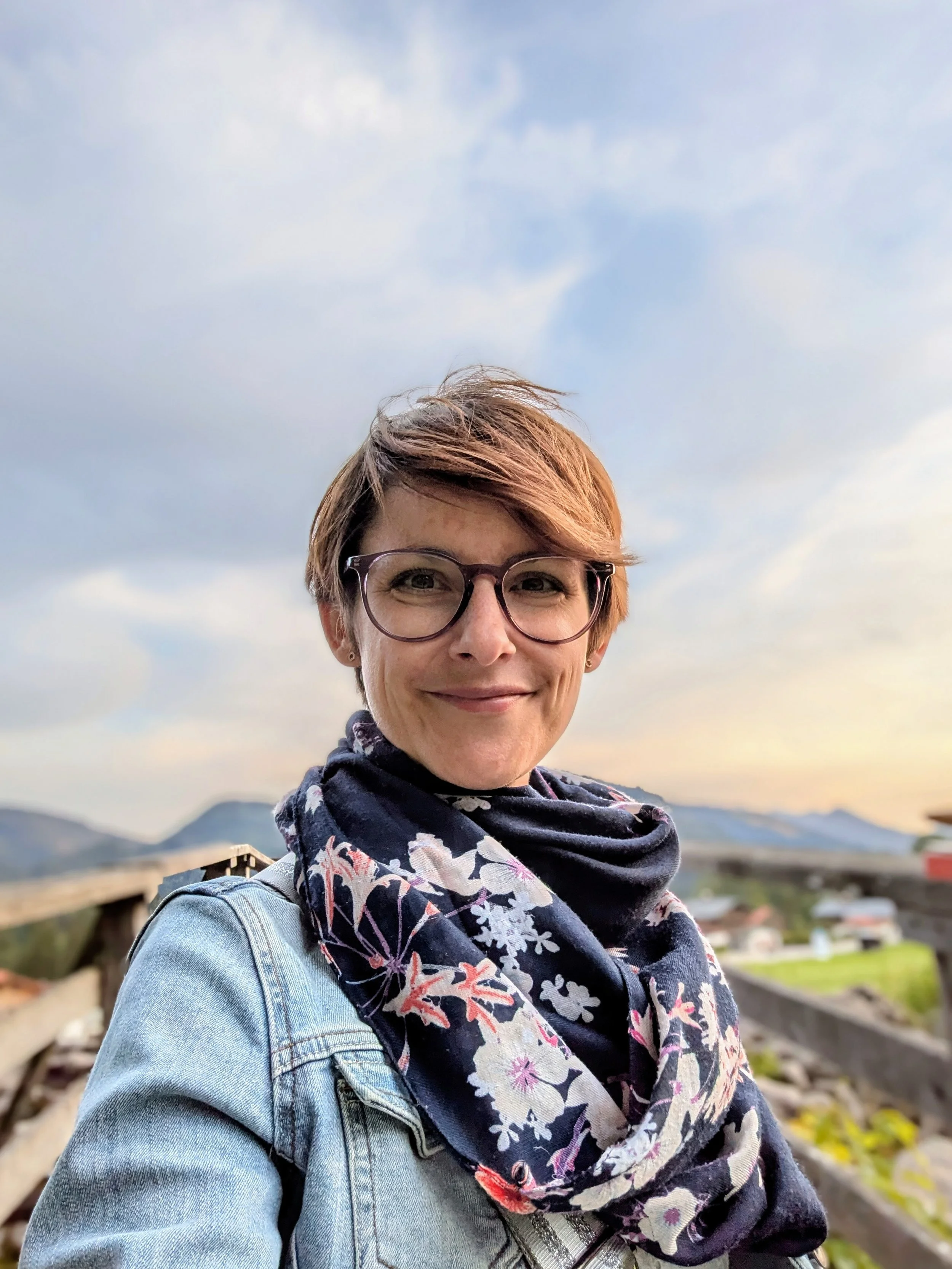 Junge Frau mit Brille, kurzer roter Haart, trägt ein Denim-Jacke und einen bunten Schal, steht im Freien mit Bergen im Hintergrund, im Sonnenuntergang.