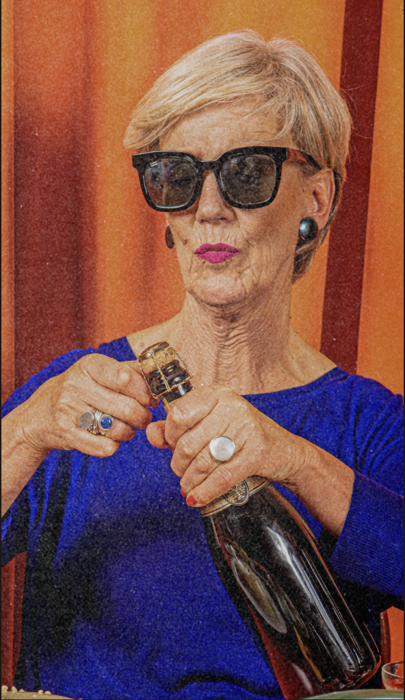 An elderly woman with short blonde hair wearing large black sunglasses, a blue top, and jewelry, opening a bottle of champagne.