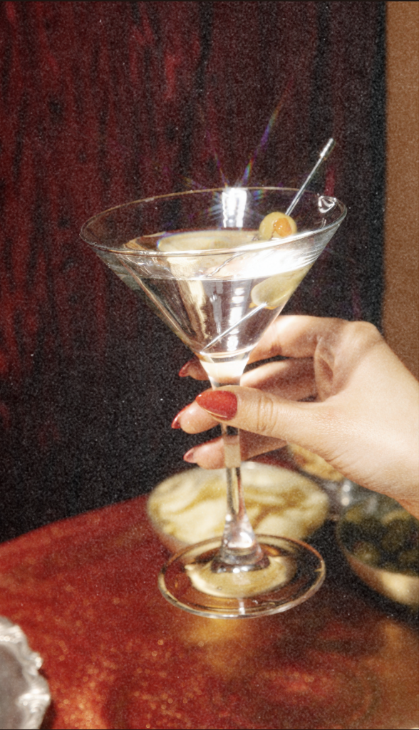 A hand with red painted nails holding a martini glass with an olive garnish and cocktail pick, set against a dark background.
