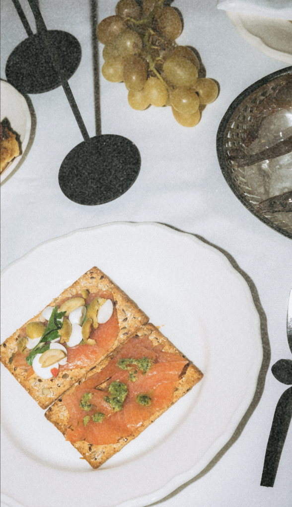 A white plate with two crispbread crackers topped with smoked salmon, cream cheese, chopped vegetables, and herbs, on a table with grapes and drinking glasses nearby.