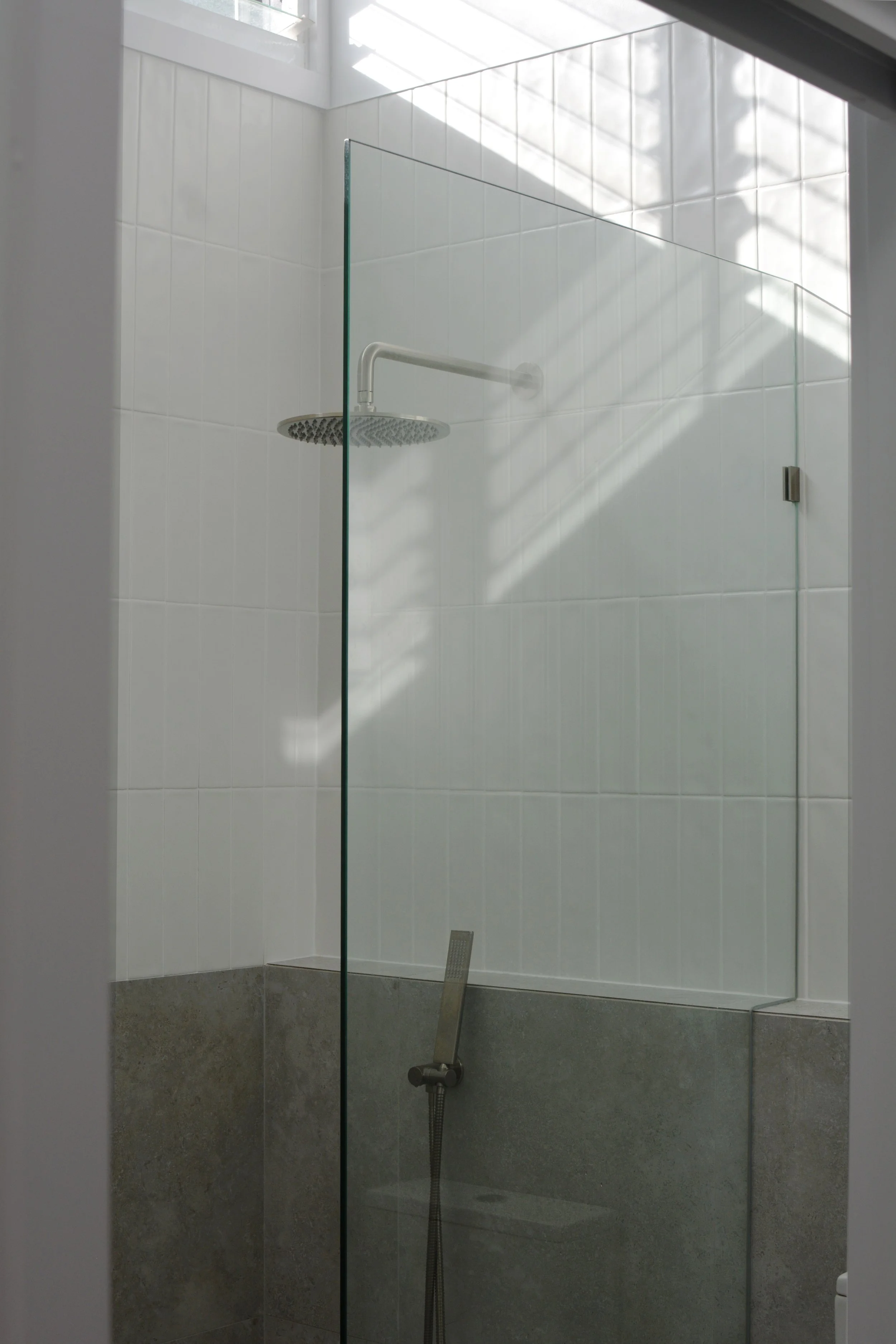 Modern bathroom shower with a glass partition, featuring a large ceiling showerhead, white tiled walls, and a wall-mounted handheld shower.