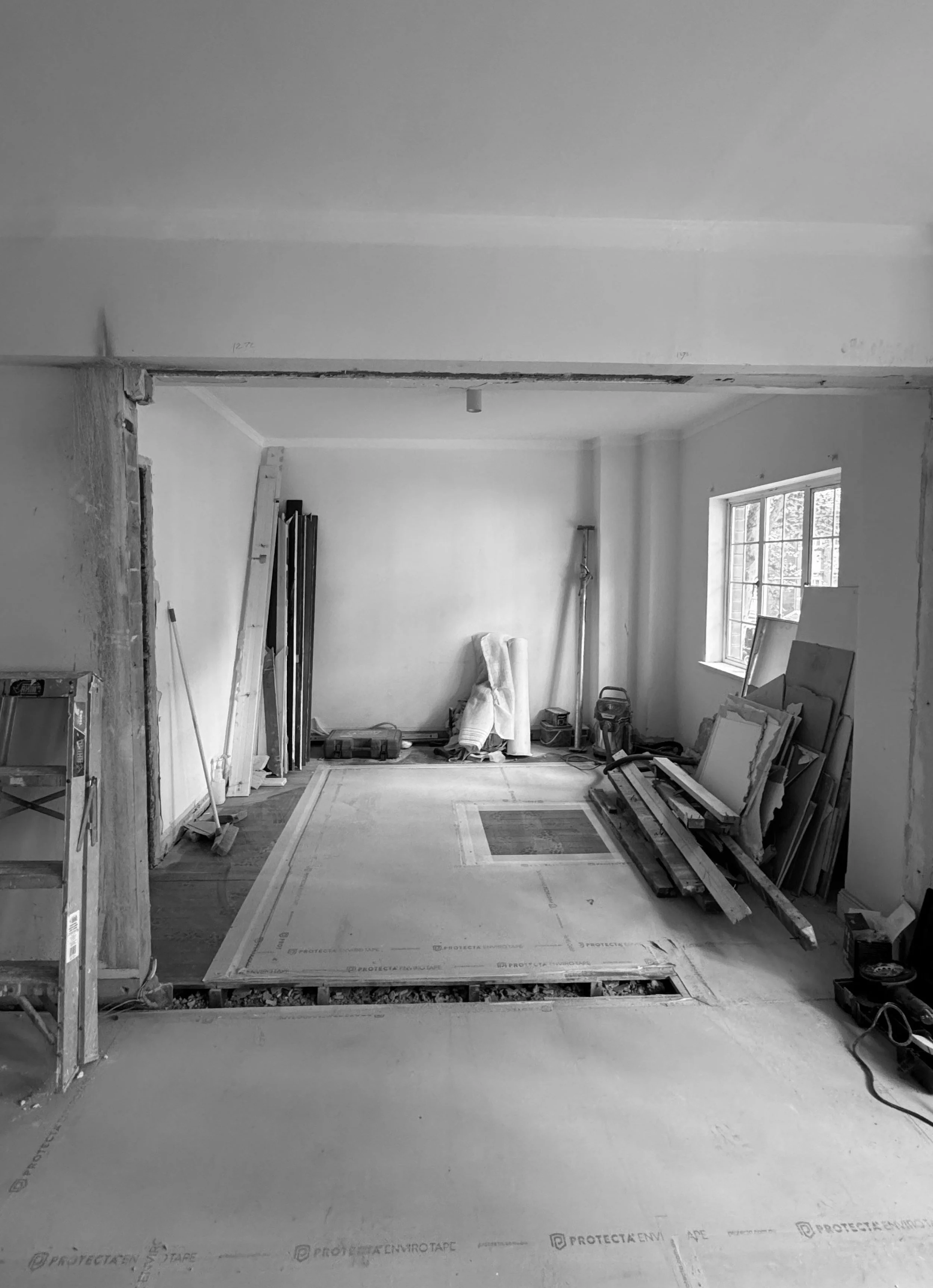 A room under construction with drywall and insulation, having construction tools, materials, and a ladder, with a window on the right side.
