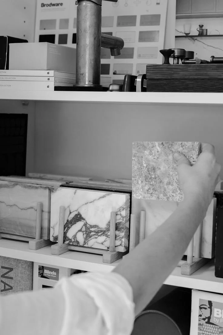 A person with a light-colored shirt holding a tile sample in front of a display of various marble and stone tile samples on shelves in a showroom or store.