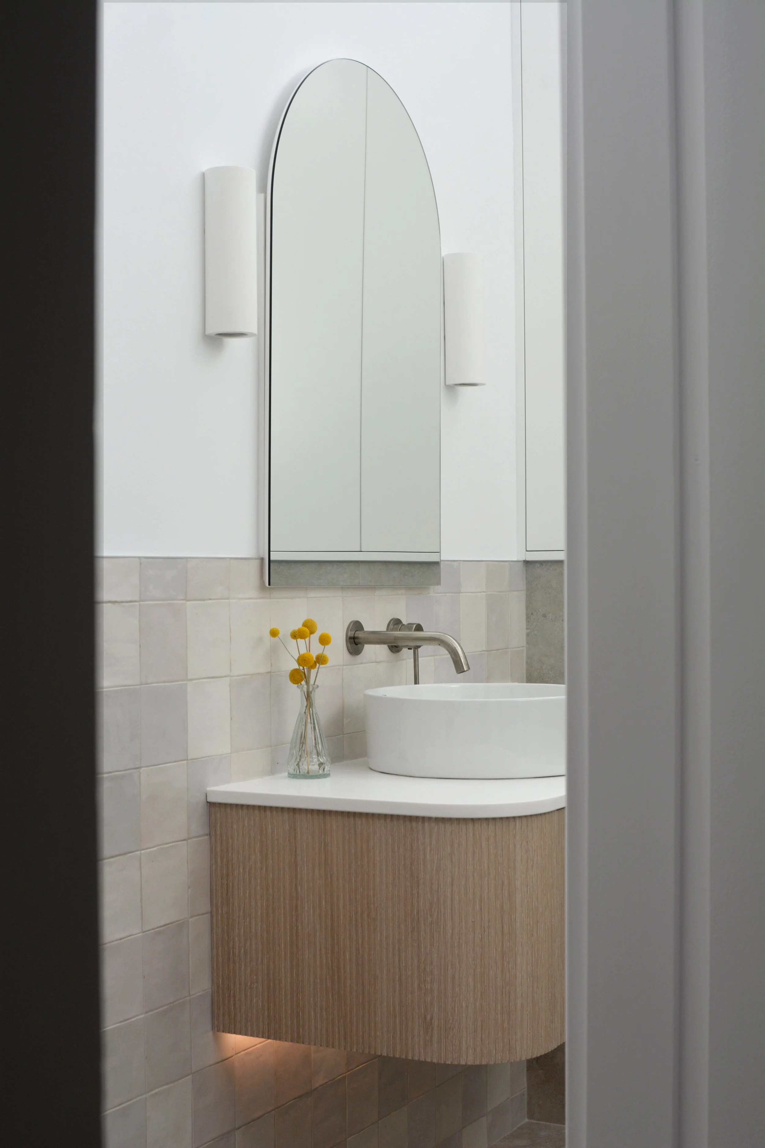 A modern bathroom with a curved wood vanity, an oval vessel sink, a silver faucet, and a decorated vase with yellow flowers. A wall-mounted mirror and white wall sconces are visible.