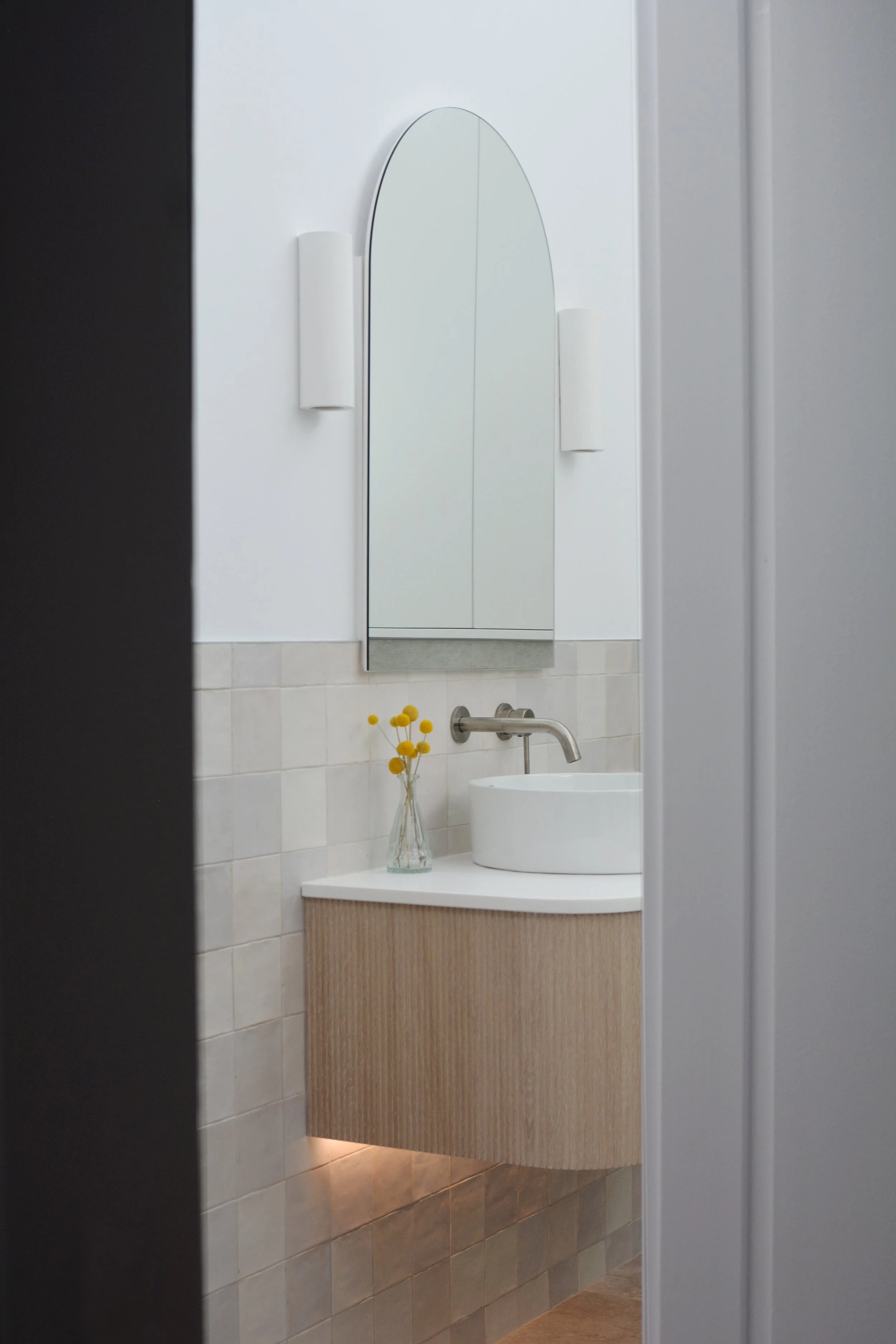 Interior view of a modern bathroom with a curved white vanity with a built-in sink, a large vertical mirror, and a bottle of yellow flowers in a glass vase.