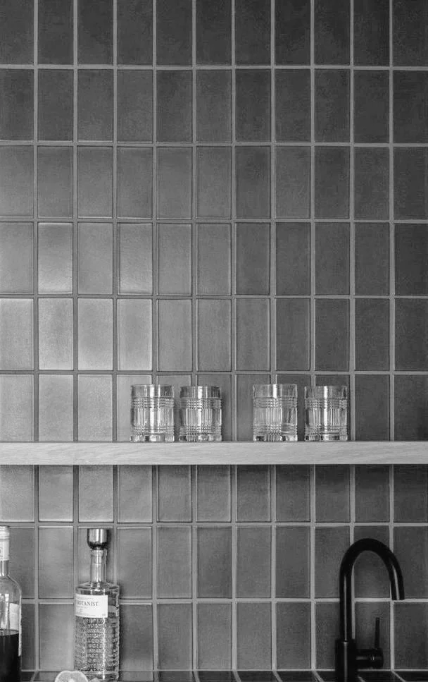 A kitchen or bar area with a tiled gray wall, a shelf with four empty glasses, and bottles of alcohol and a black faucet below the shelf.