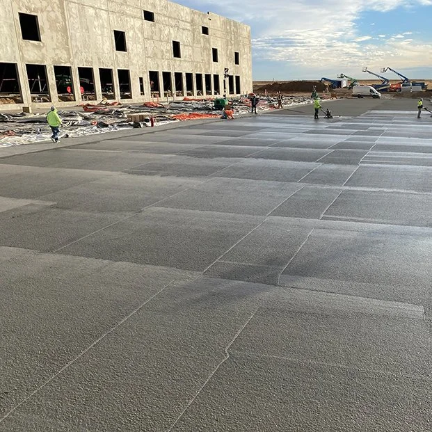 jscreed Construction workers on a building site installing large gray tiles, with a partially built concrete structure and construction equipment in the background.