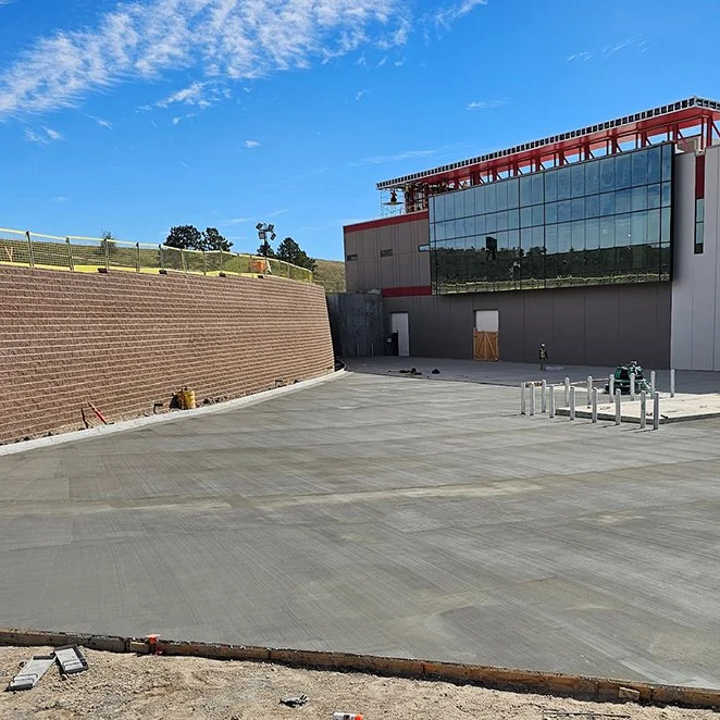 jscreed Construction site with a newly poured concrete area, a brick retaining wall on the left, and a modern building with reflective glass windows on the right under a clear blue sky Colorado