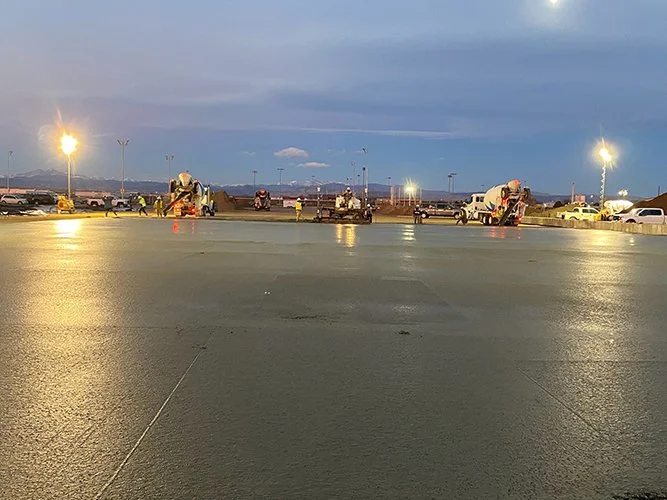 A wet parking lot at dusk with jscreed workers and equipment, and a city skyline in the background.