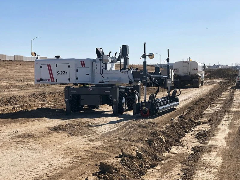 jscreed construction site with a robotic construction vehicle and a water tanker truck on a dirt road under a clear blue sky