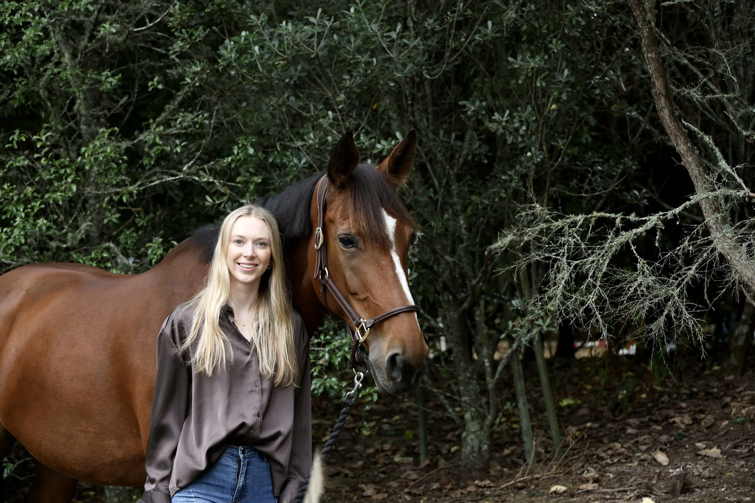 A woman with long blonde hair standing next to a brown horse with a white stripe on its face, in front of green foliage.