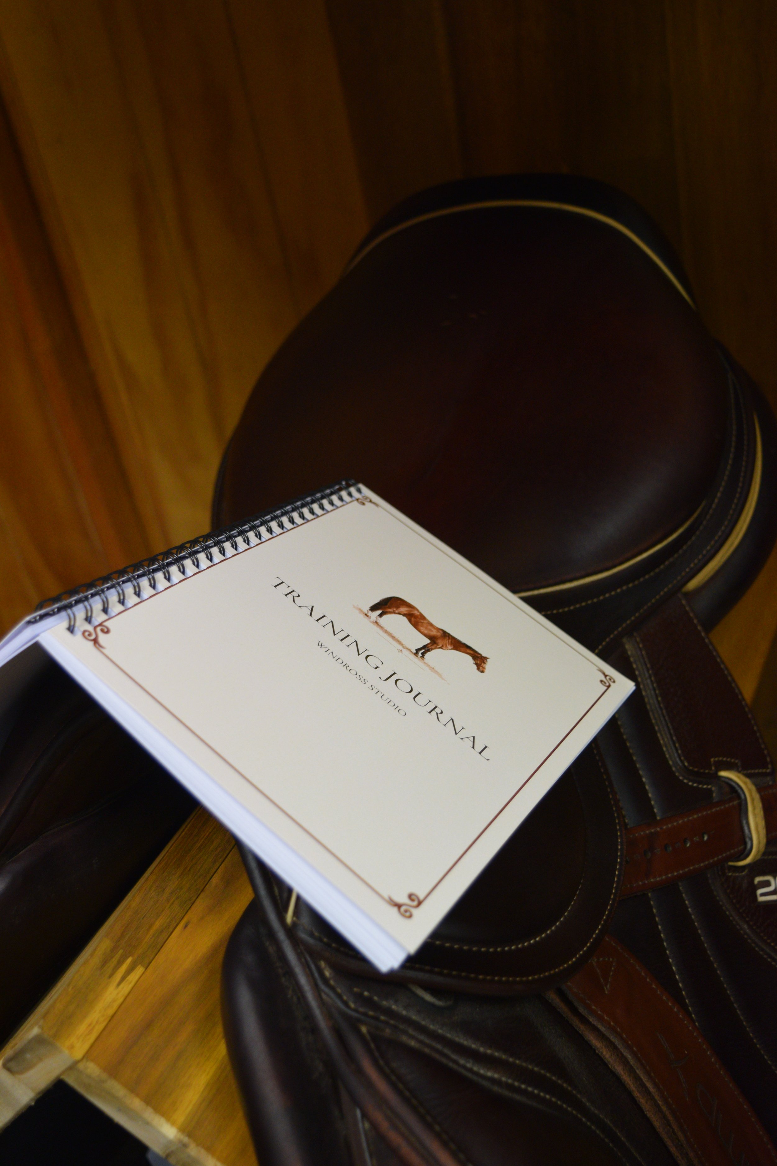 Book titled 'Training Journal' resting on a saddle with a wooden wall in the background.