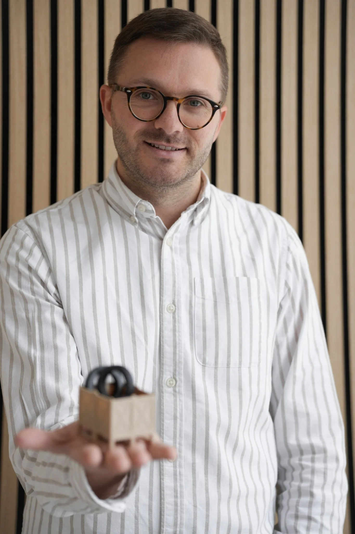 Ein Mann in einer weißen gestreiften Hemd trägt eine Brille und hält eine kleine Pappbox mit einem schwarzen Objekt darin in der Hand, im Hintergrund eine Wand mit vertikalen Holzlamellen.