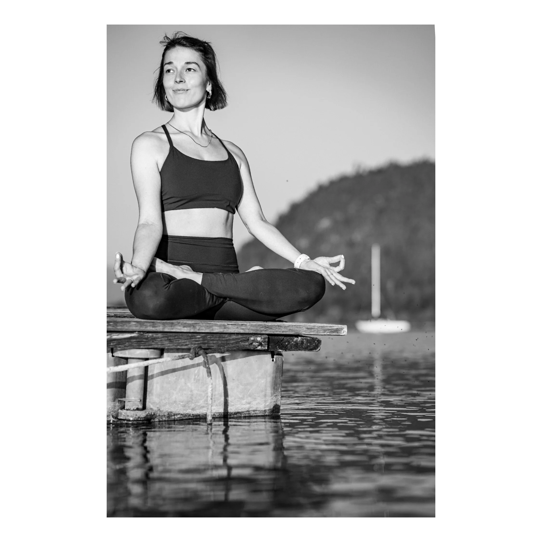 Schwarz-Weiß-Foto einer jungen Yoga-Lehrerin, die auf einer Cloud-Dekoration im Wasser Meditationshaltung hält. Im Hintergrund ist ein blauer Himmel und Wasser. Die Frau trägt eine Sport-BH und legere Kleidung, wirkt entspannt und konzentriert.