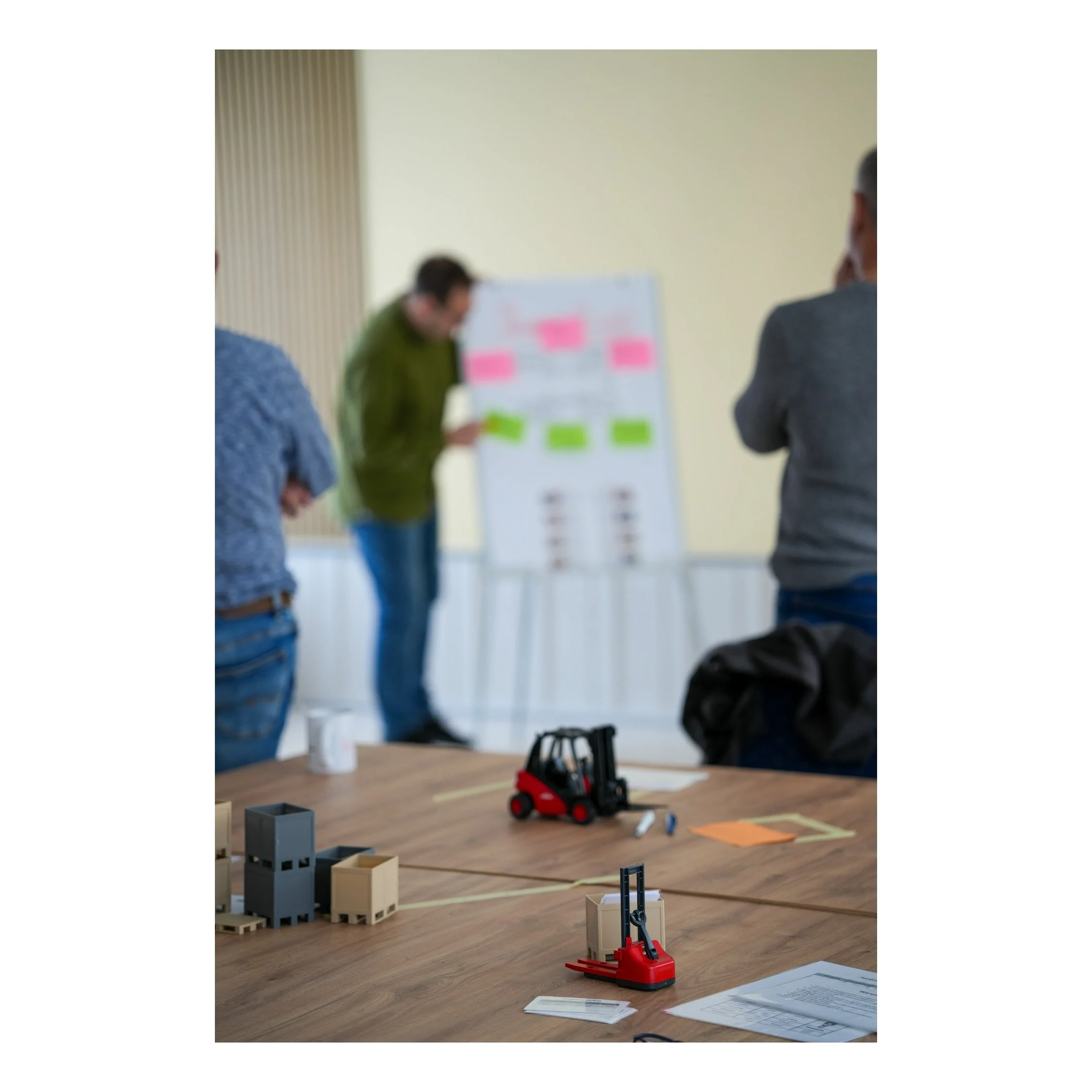 Shooting - Business Reportage eines Startup’s im Bereich Logistikberatung. Workshop mit Modellen, Werkzeugen und Dokumenten auf einem Holztisch, im Hintergrund Personen bei einer Präsentation oder Diskussion.
