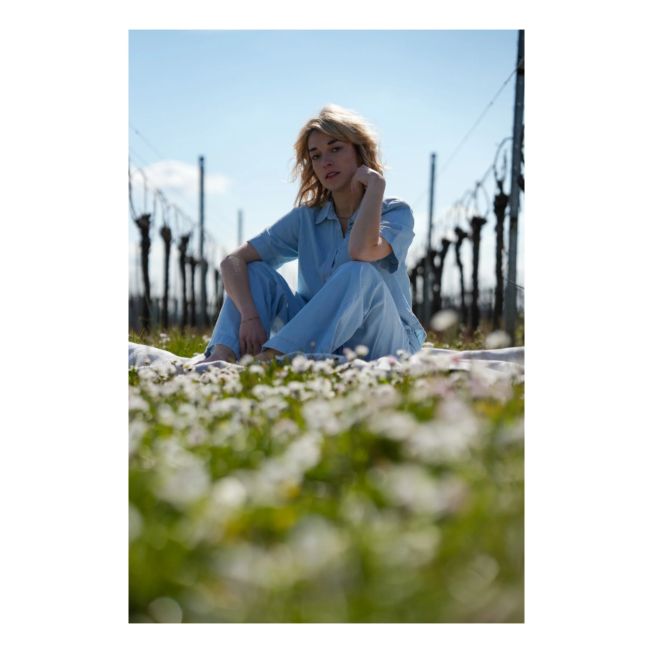 Shooting Portrait Outdoor in Farbe. Eine junge Frau sitzt im Freien auf einer Wiese, umgeben von blühenden Blumen, bei Tageslicht. Sie hat blondes, leicht gewelltes Haar und trägt eine hellblaue Hose und ein Hemd.