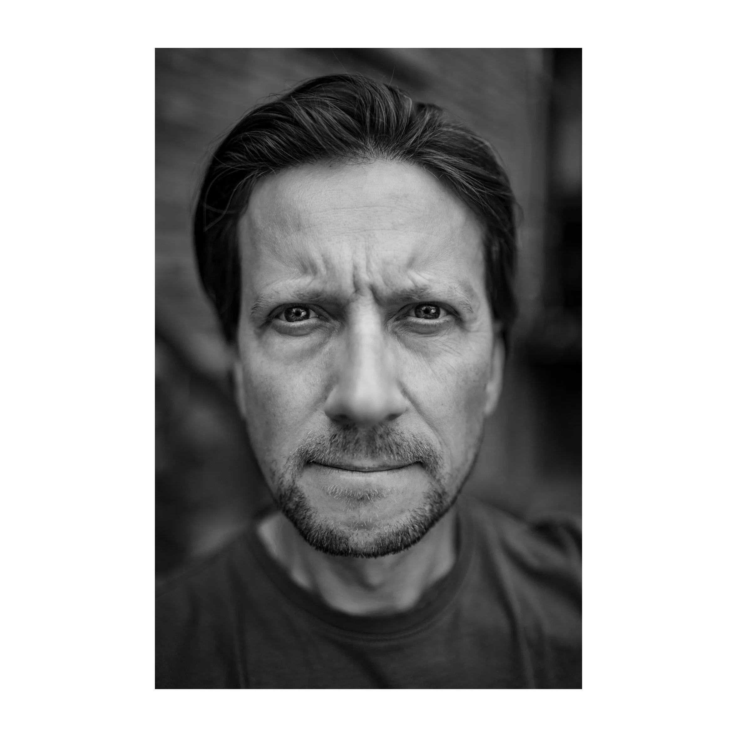 Schwarz-weiß Porträt eines Mannes mit langen Haaren, blauen Augen, einem leichten Bart, der direkt in die Kamera blickt.